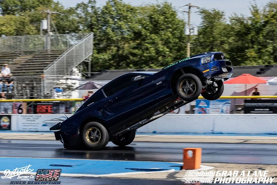 A dark blue Mustang lifting its front wheels off the line at the drag strip